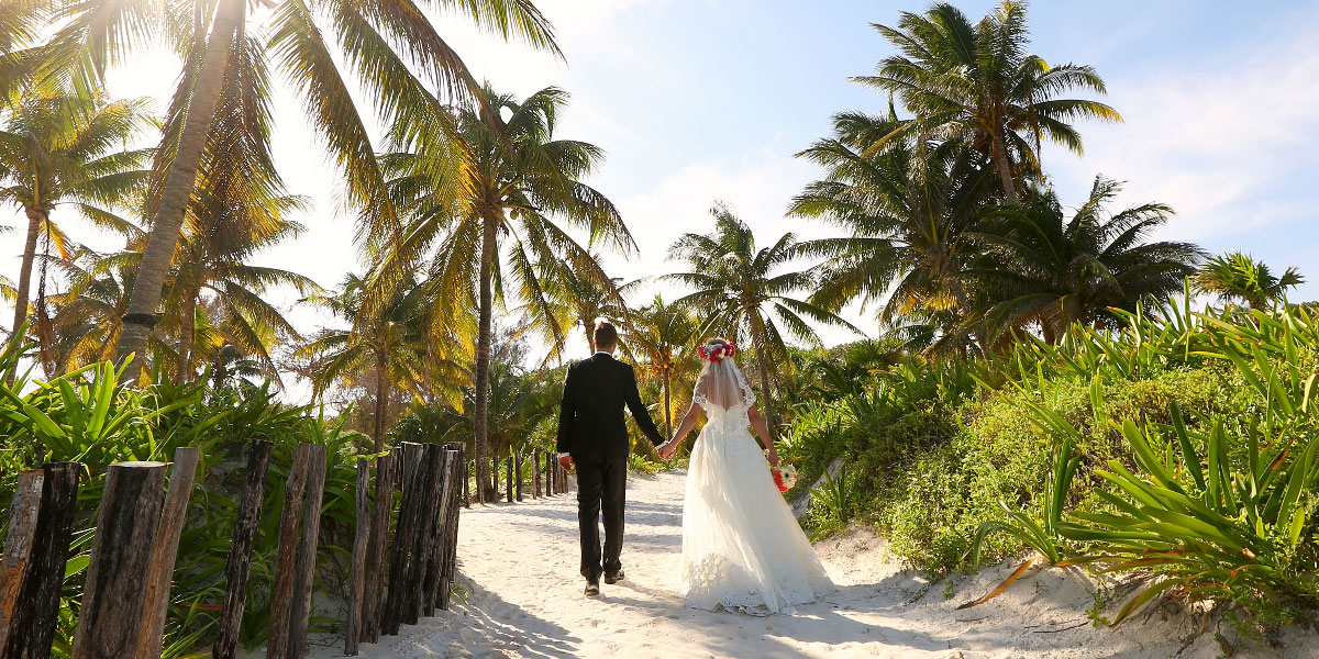 Bride and groom walking down sand path to beach.